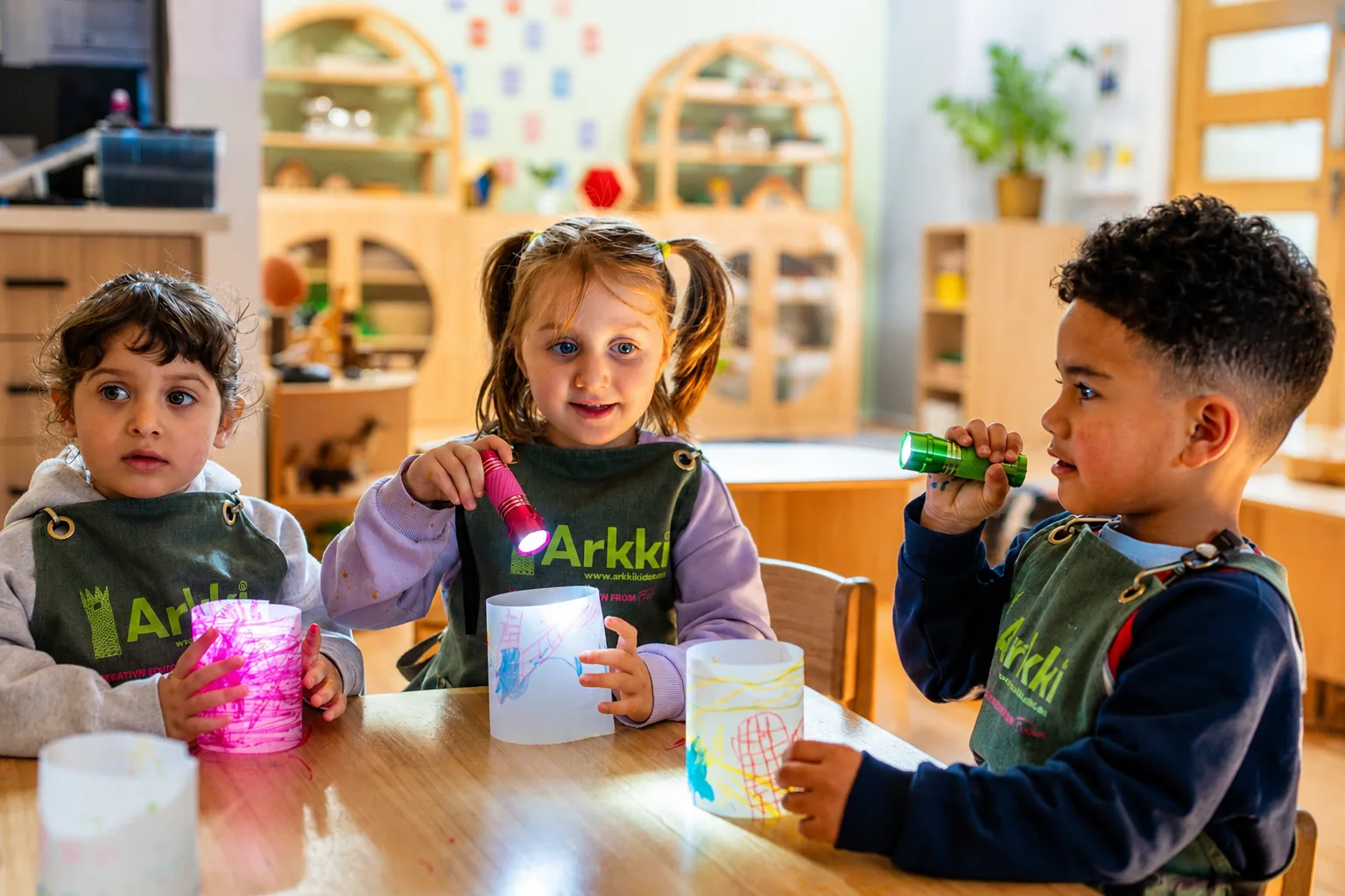 Three children in Arkki aprons at a table, shining torches into the paper lanterns they have drawn on